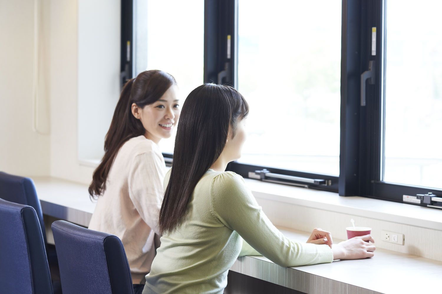 Dos personas sentadas a una mesa junto a una ventana en una habitación de diseño sencillo, con vistas al exterior.