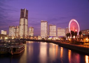 A cityscape at night with illuminated skyscrapers and a large Ferris wheel reflecting on a calm river. - Sotetsu Fresa Inn Yokohama Totsuka (Yokohama)
