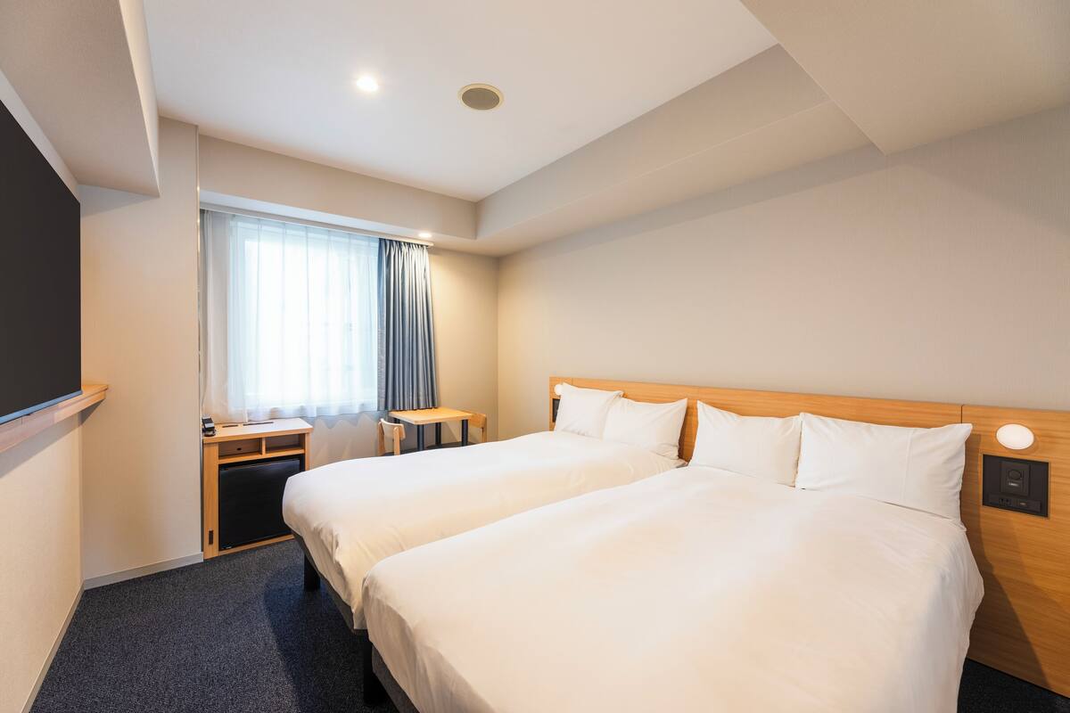Een hotelkamer met twee bedden, een bureau, een stoel, een raam met gordijnen en een bedieningspaneel aan de muur.