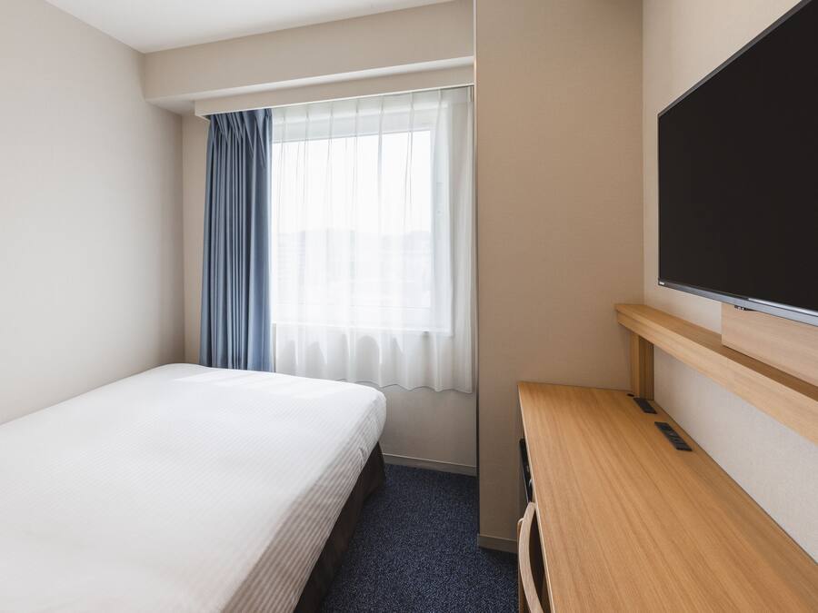 A hotel room with a single bed, a desk, a television, and a window with curtains.
