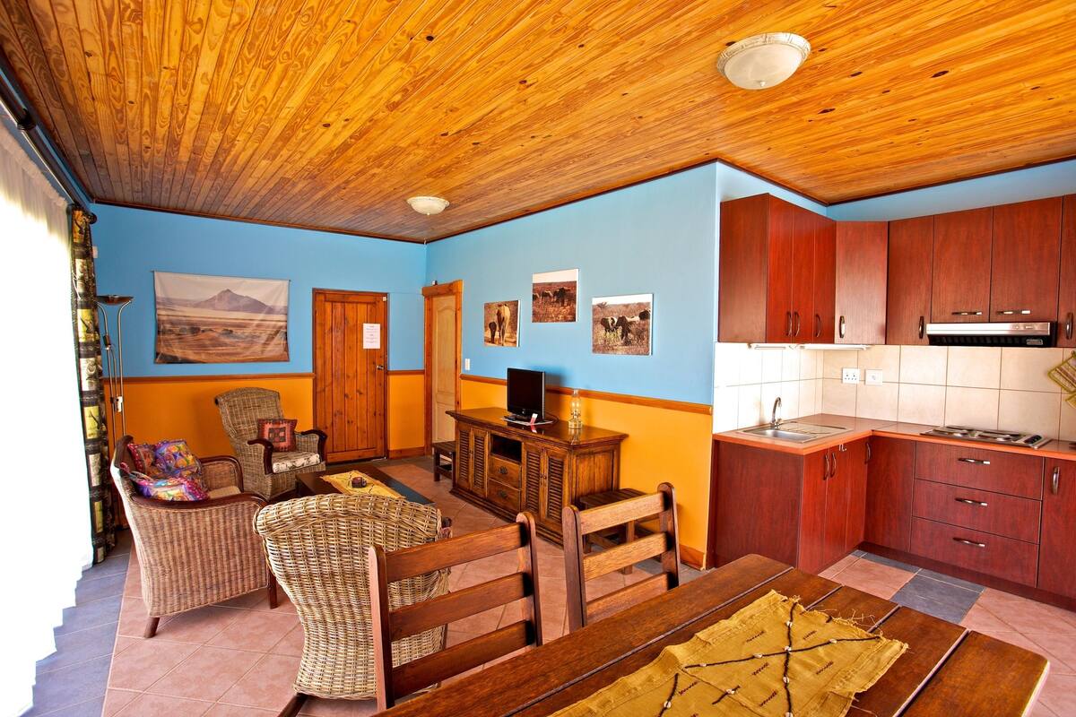 Ein Wohnzimmer mit Korbmöbeln, einem Esstisch und einer Küchenzeile mit Holzschränken und einer Spüle.