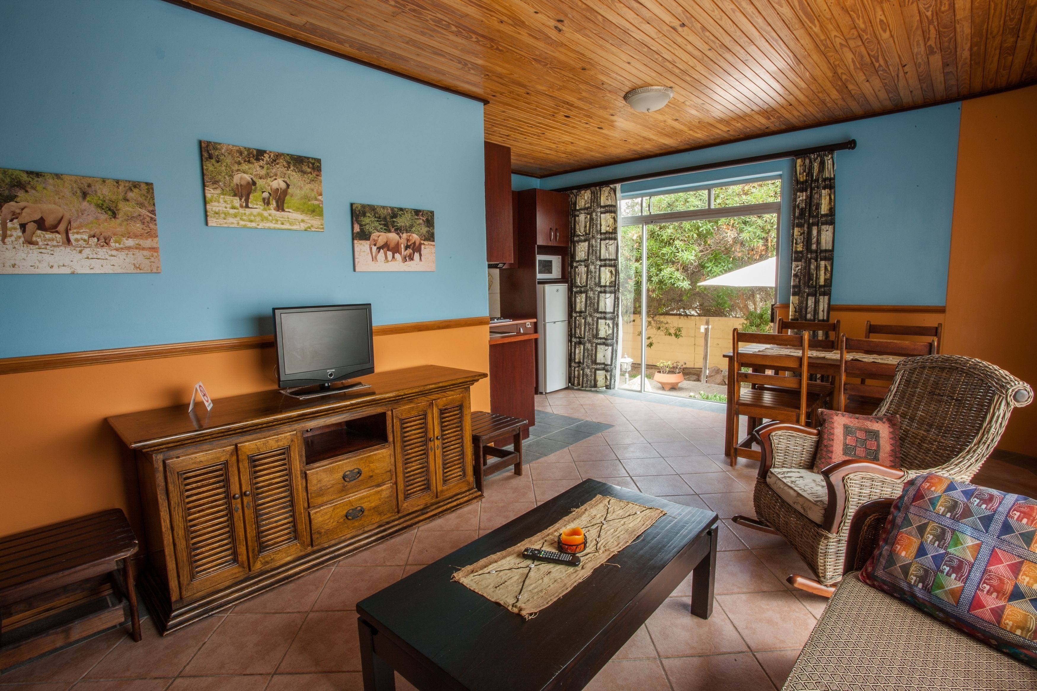 Una habitación con techo de madera, un televisor sobre un mueble, un comedor con mobiliario de mimbre y vistas a través de una puerta de cristal.