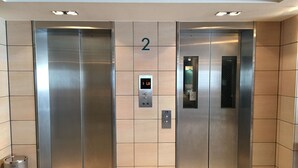 Zwei Aufzugtüren mit digitaler Anzeige und Tastenfeld, sowie ein Mülleimer links davon.