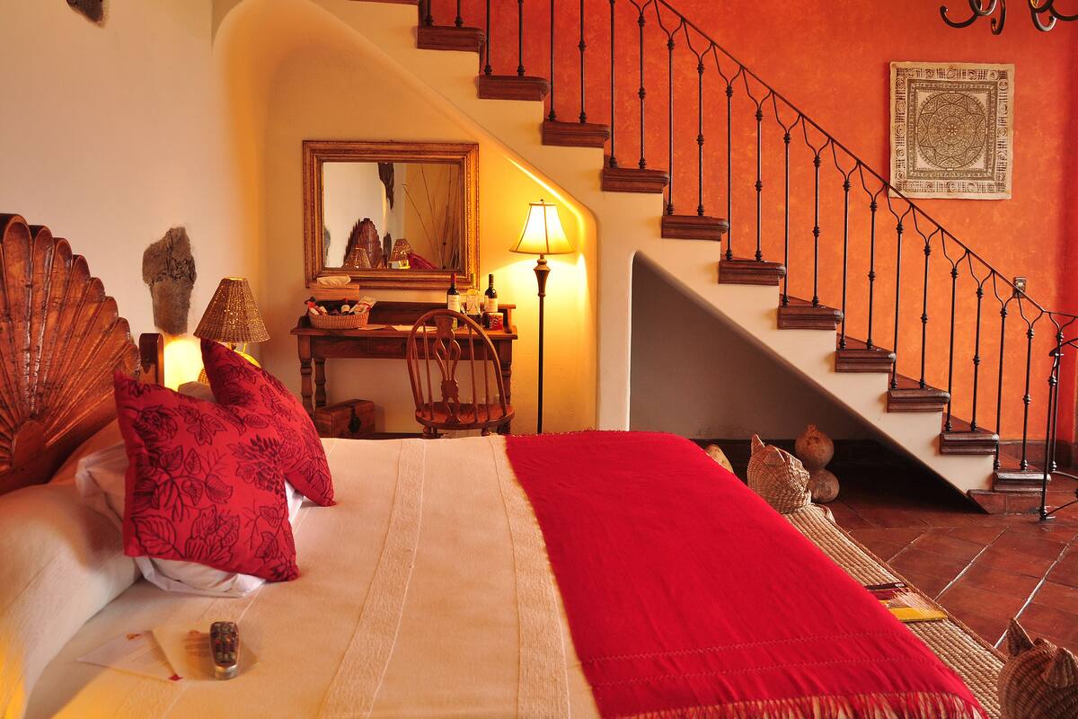 A bedroom with a large bed, a desk with a chair, a staircase, and a chandelier.