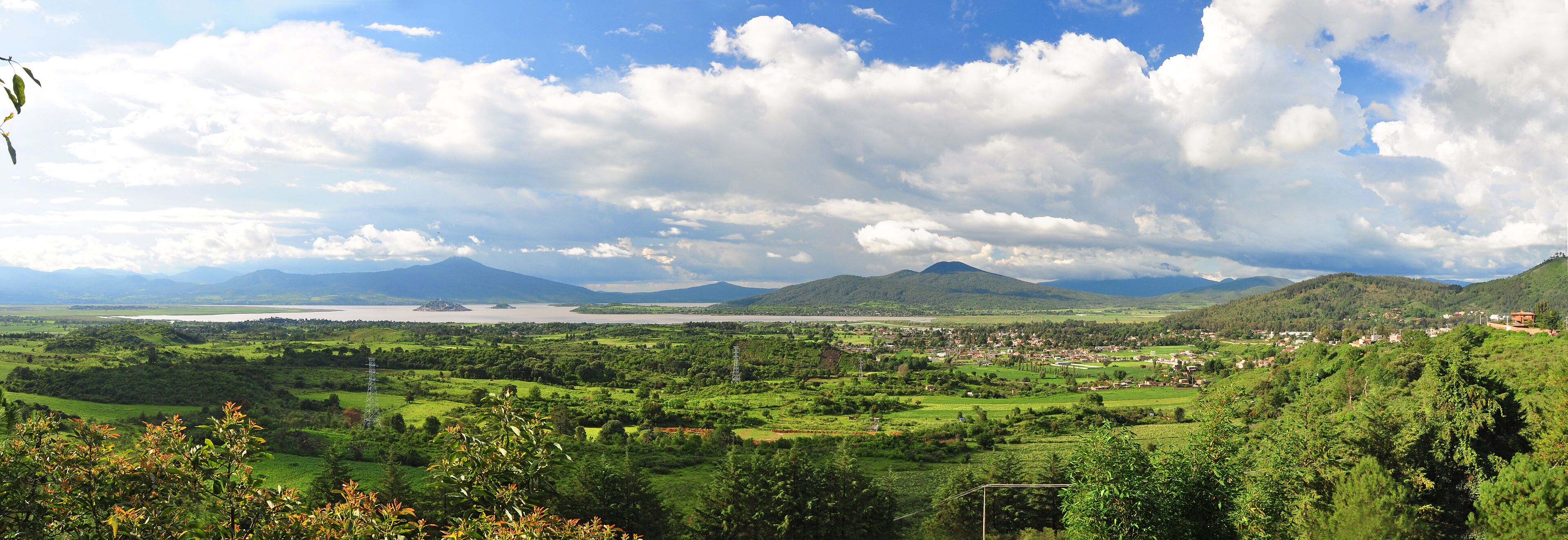 Vista panorámica de un paisaje verde exuberante con un pueblo en el valle, montañas al fondo y una masa de agua.