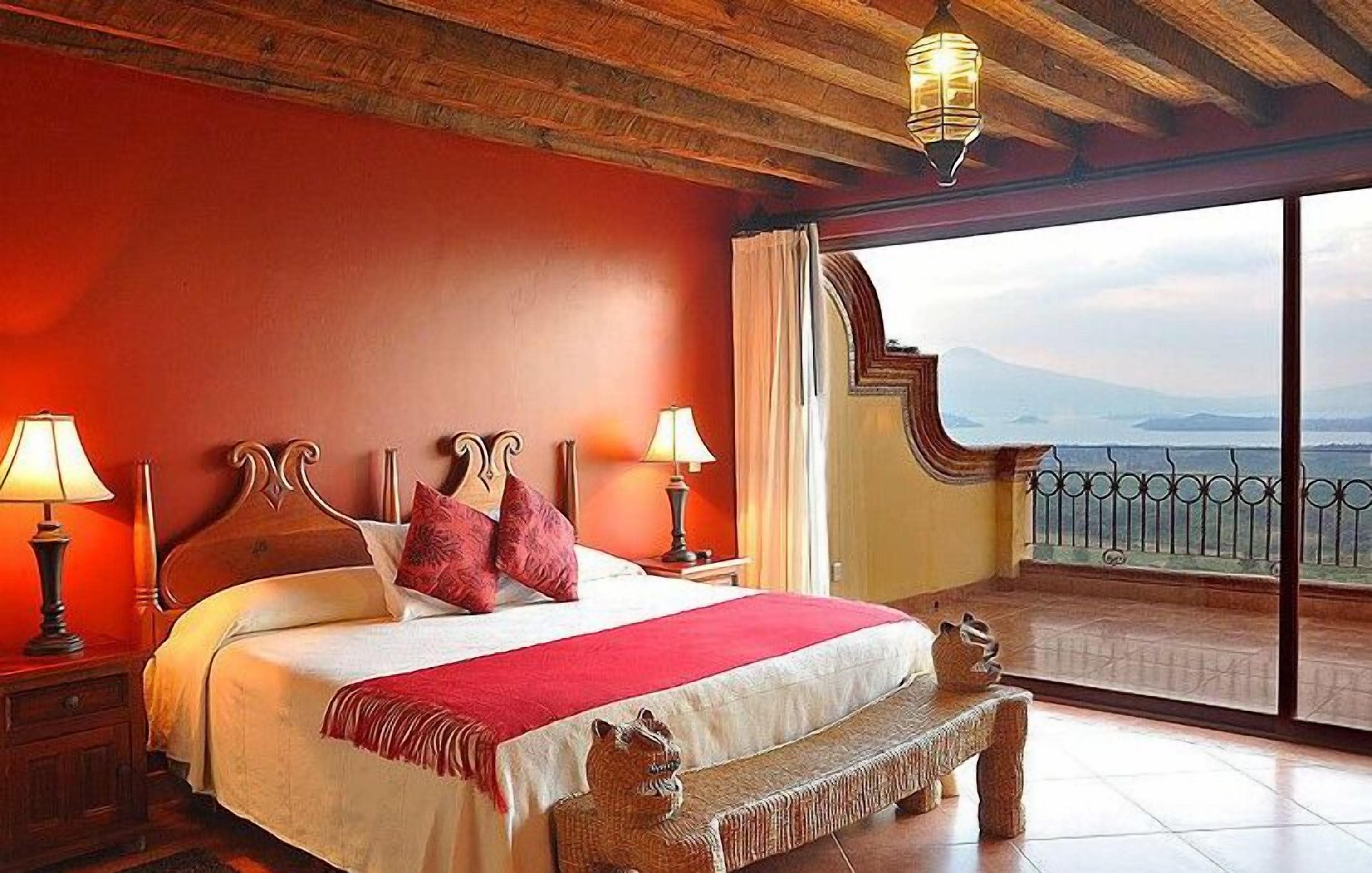 Une chambre avec un grand lit, une tête de lit en bois, deux lampes de chevet, un banc et une vue sur la mer depuis un balcon.