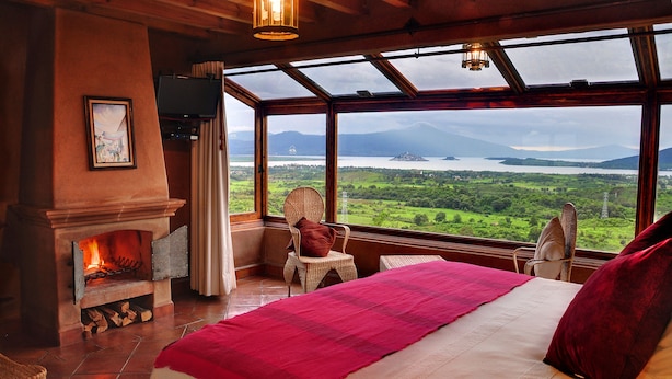 A bedroom with a large bed, a fireplace, wicker chairs, and a view of a lake and mountains. - Hotel Porton Del Cielo (Patzcuaro)