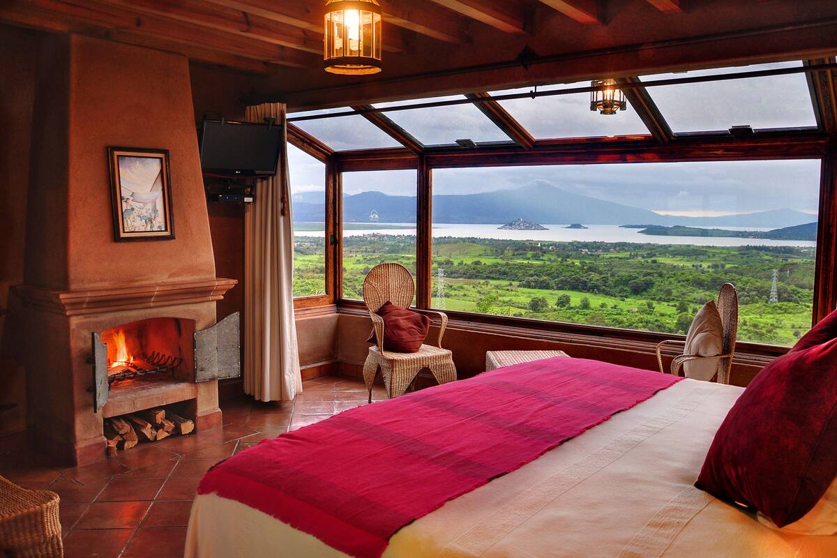 A bedroom with a large bed, a fireplace, wicker chairs, and a view of a lake and mountains.
