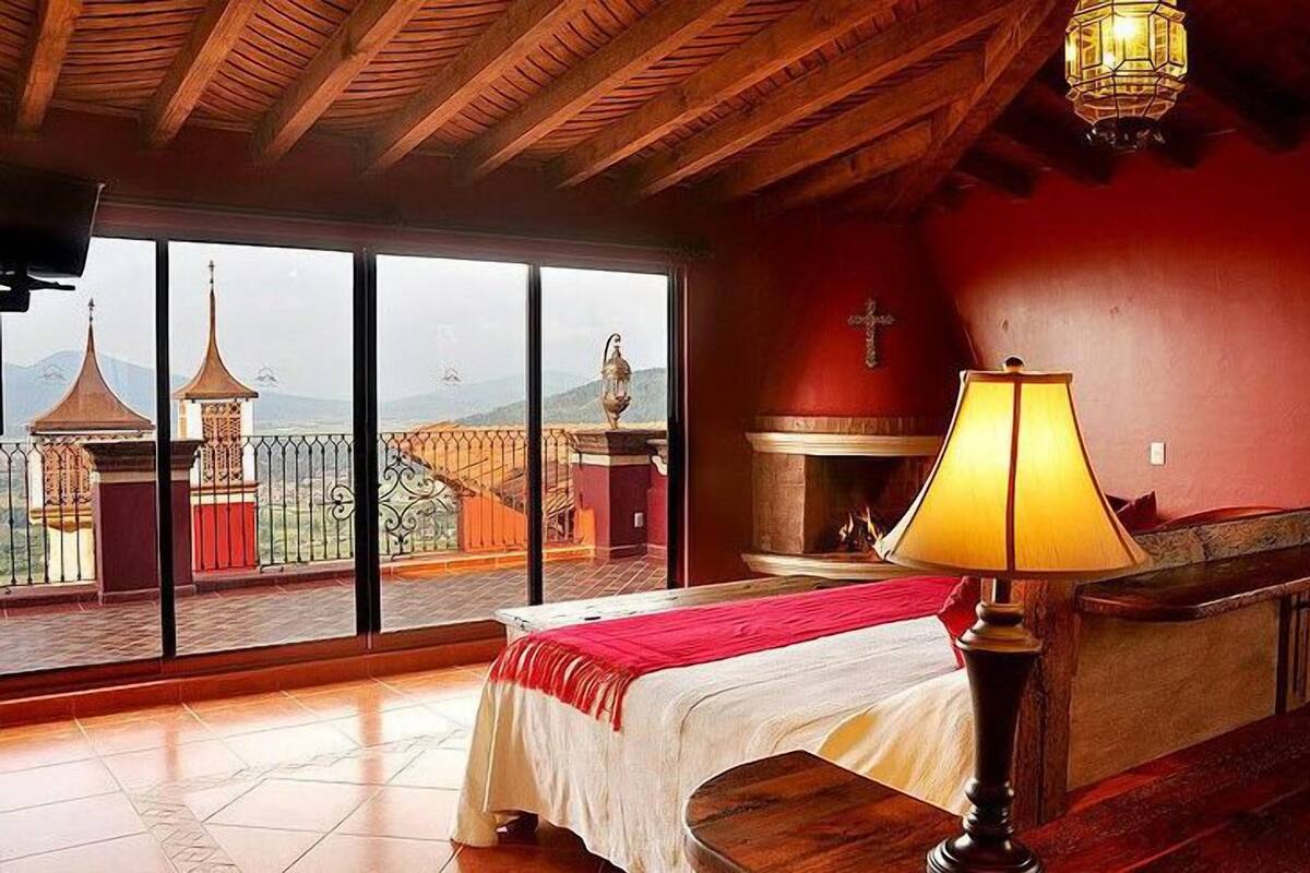 A room with a bed, a lamp, a fireplace, and a view of buildings through large windows.