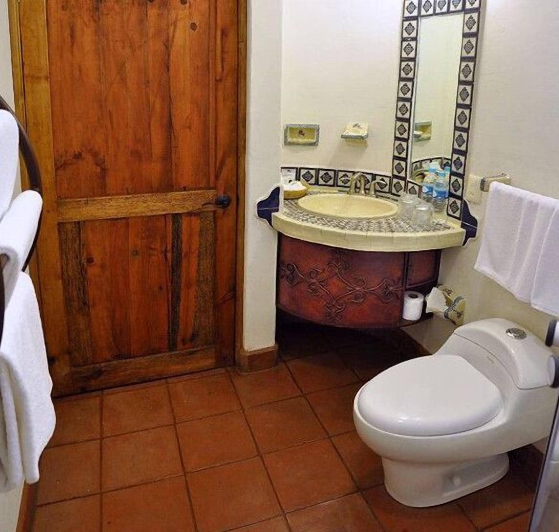 A bathroom with a wooden door, a sink with a decorative basin, a toilet, and a mirror with a patterned frame.