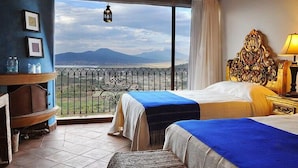 A hotel room with two beds, a balcony with a view of mountains, a wooden desk, and a decorative headboard. - Hotel Porton Del Cielo (Patzcuaro)