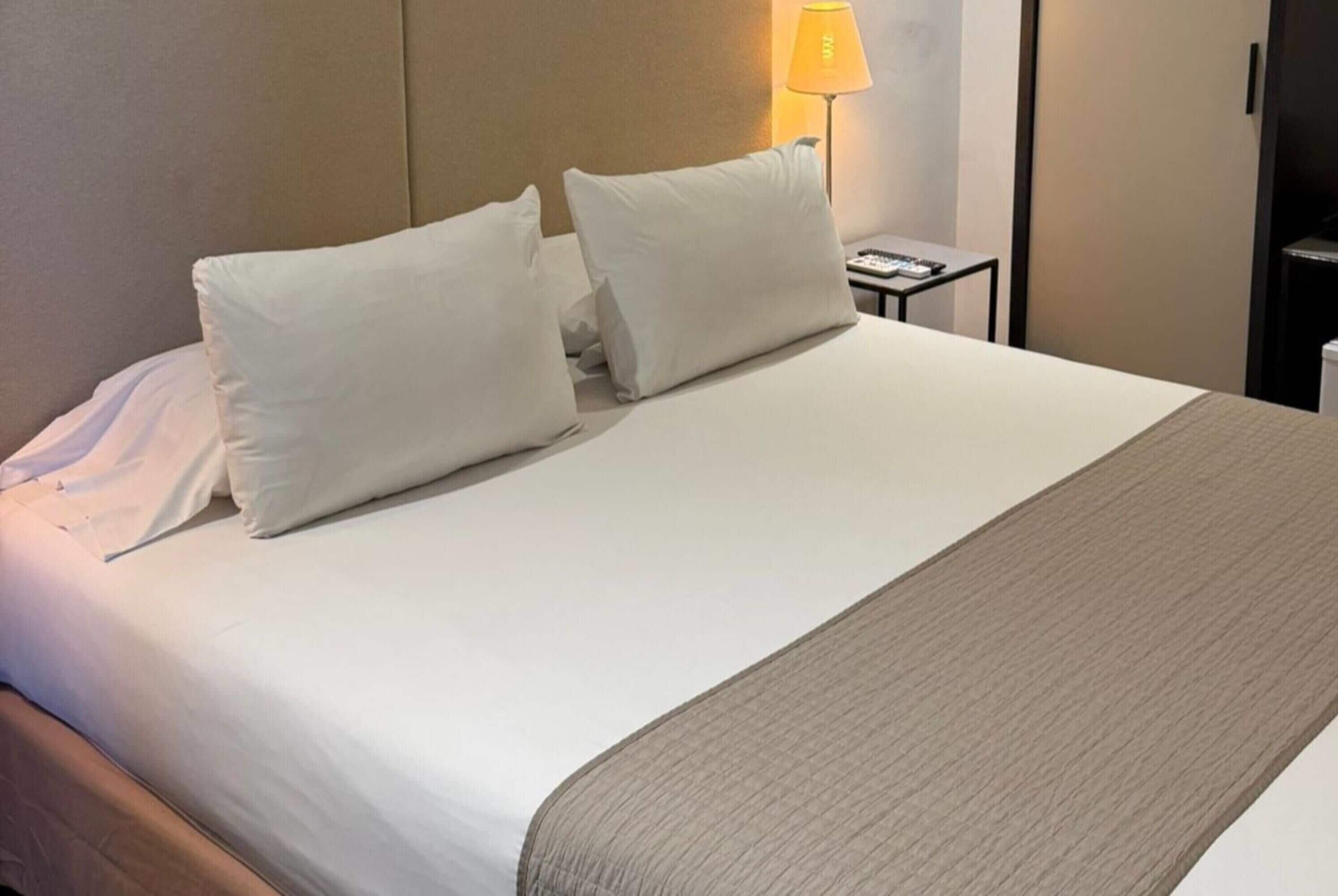 Un lit bien fait, avec du linge de lit blanc et deux oreillers, une table de chevet avec une lampe et une tête de lit en bois.