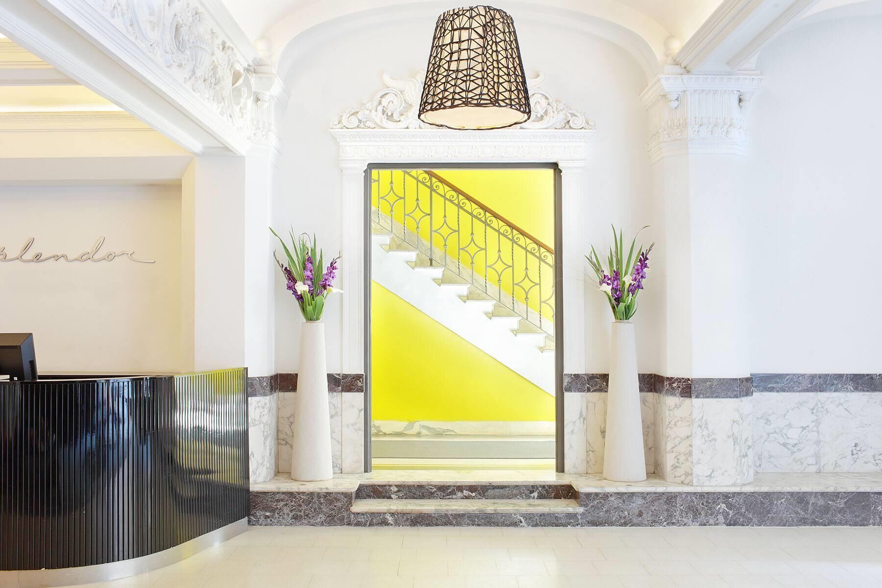 A reception area with a marble floor, a reception desk, a staircase with a yellow wall, and two vases with purple flowers.