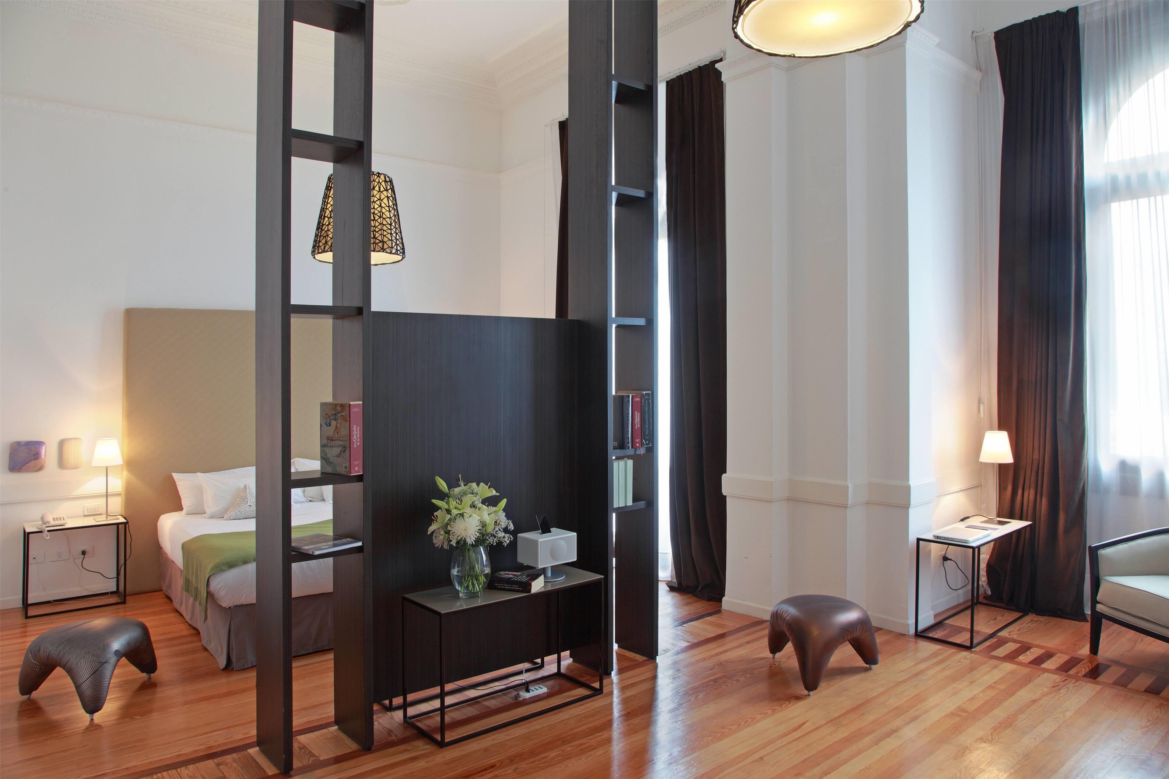 A modern hotel room with a bed, bedside table, lamp, and a small stool.
