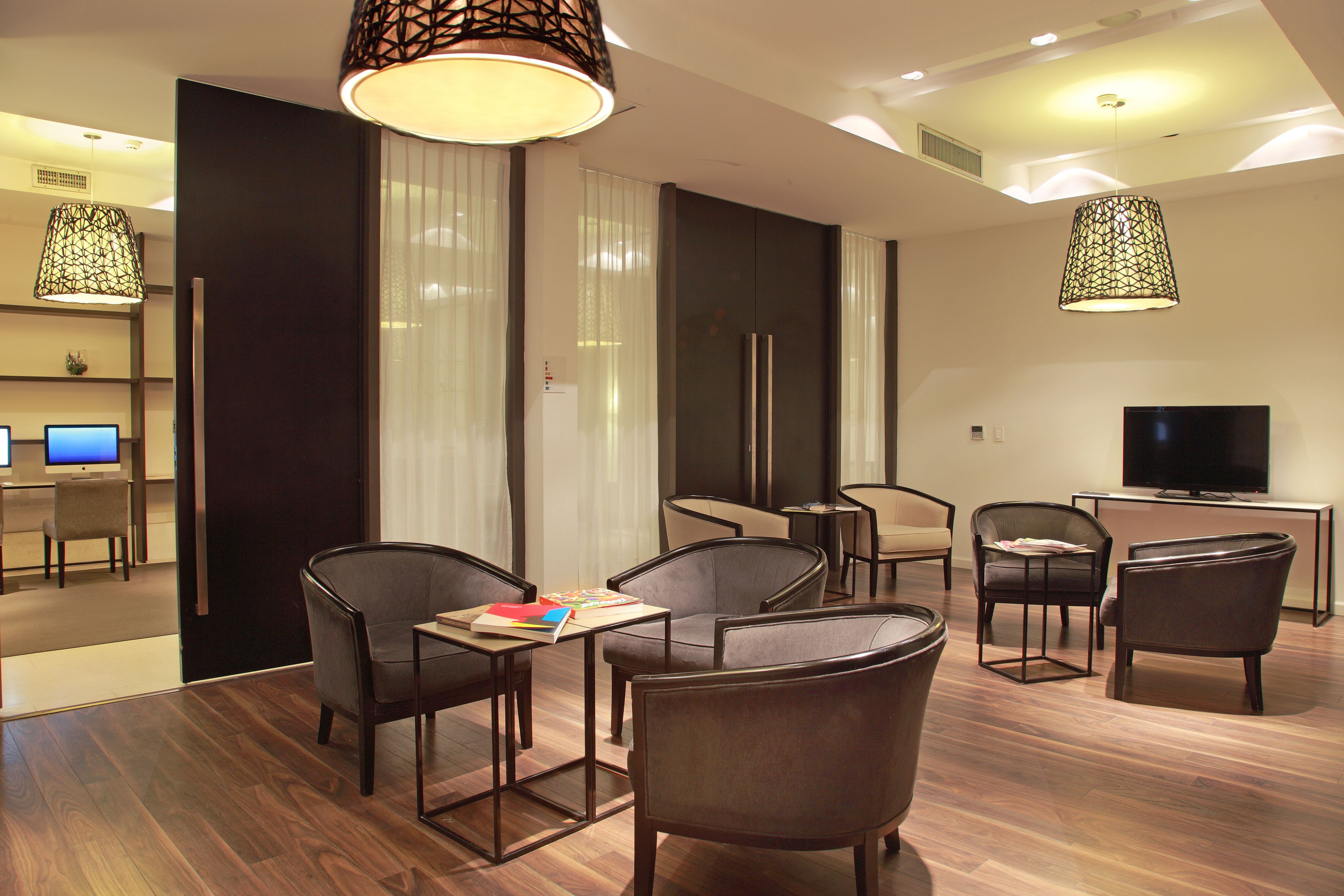 A modern hotel room with a wooden floor, a dining area with chairs and a table, a TV, and a large window with curtains.