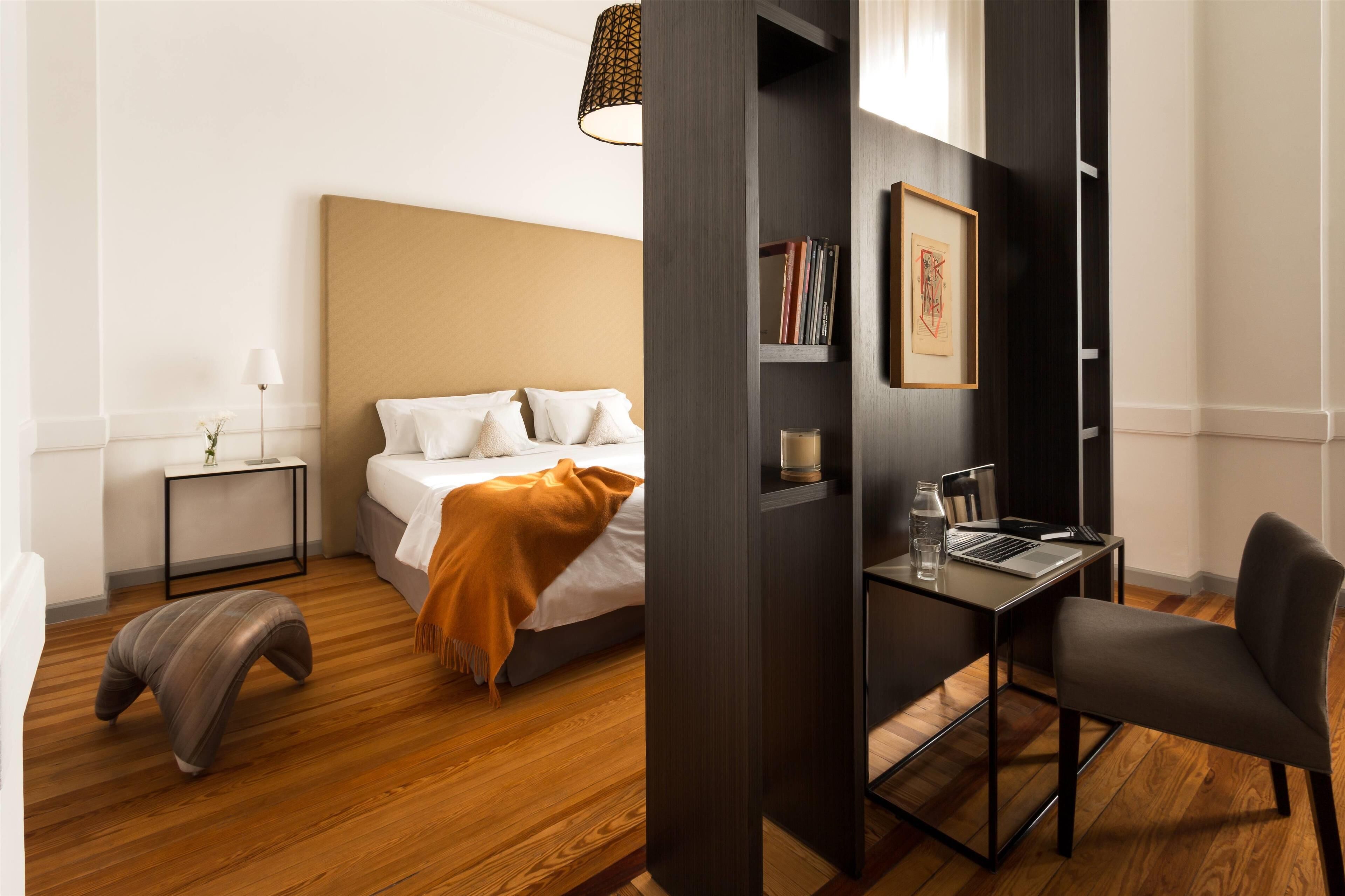 A modern hotel room with a bed, bedside table, lamp, and a desk with a chair.