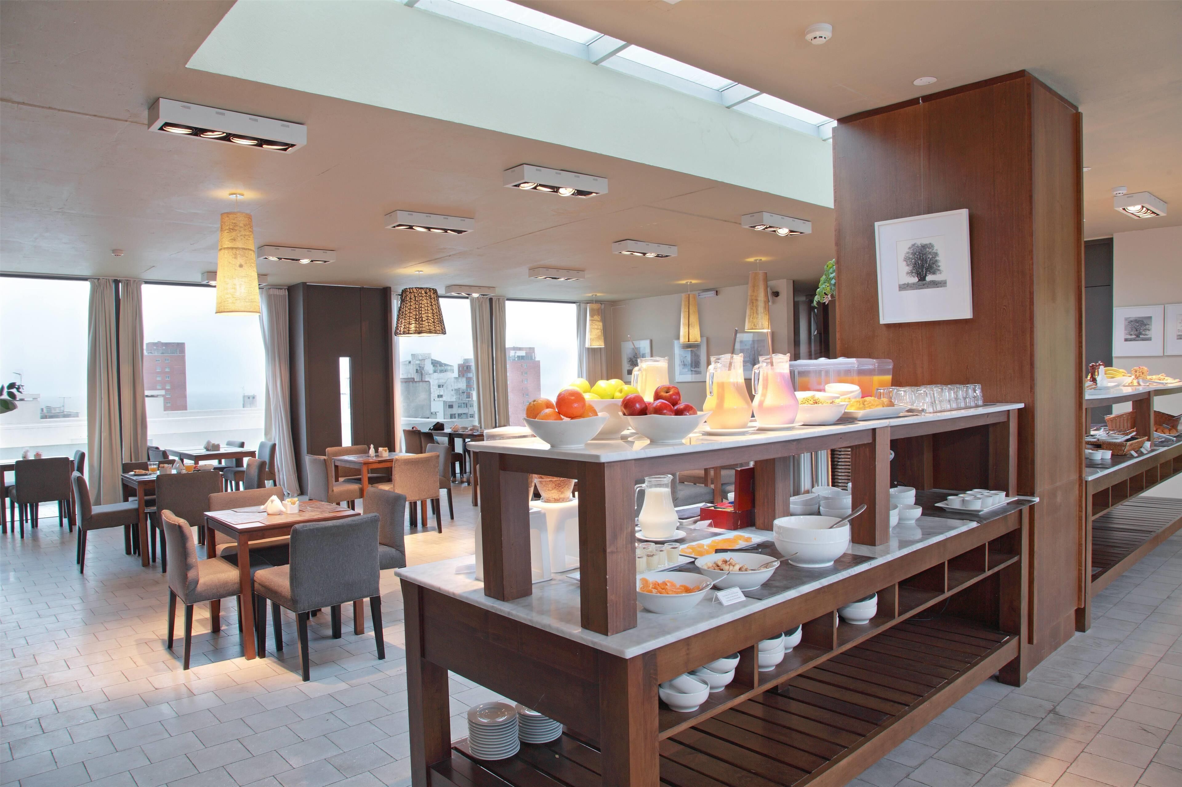 A modern hotel dining area with a long buffet table, multiple seating arrangements, and large windows offering city views.