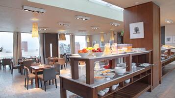 A modern hotel dining area with a long buffet table, multiple seating arrangements, and large windows offering city views.