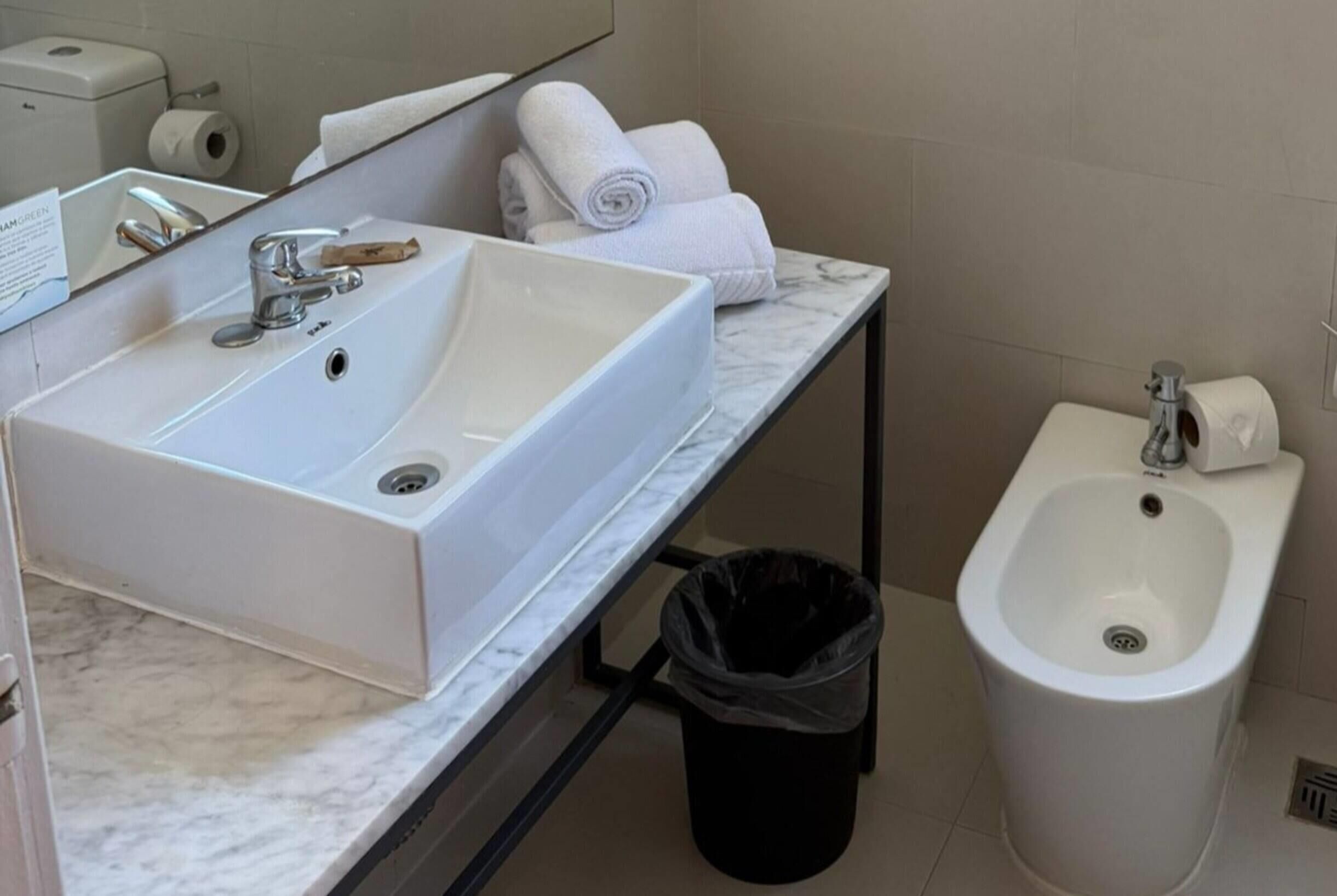 A bathroom with a white sink, a bidet, a black trash bin, and a towel rack with folded towels.