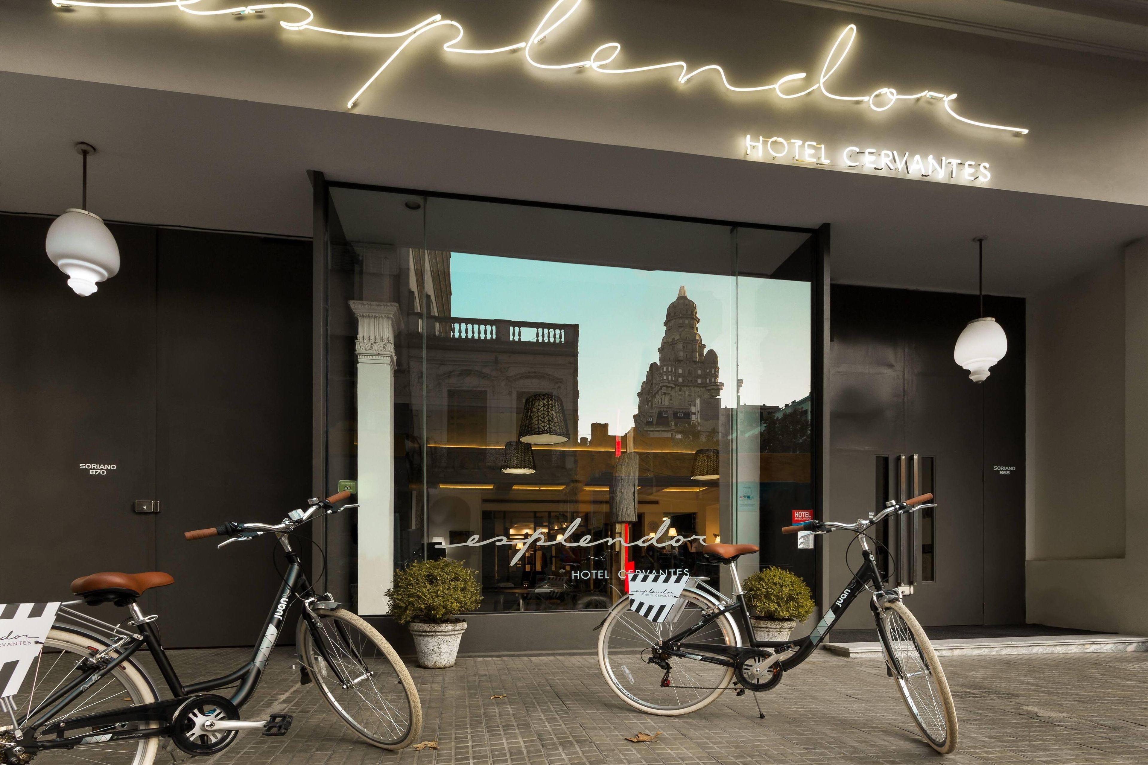 A hotel entrance with a neon sign, two bicycles parked in front, and a view of a historic building through the window.