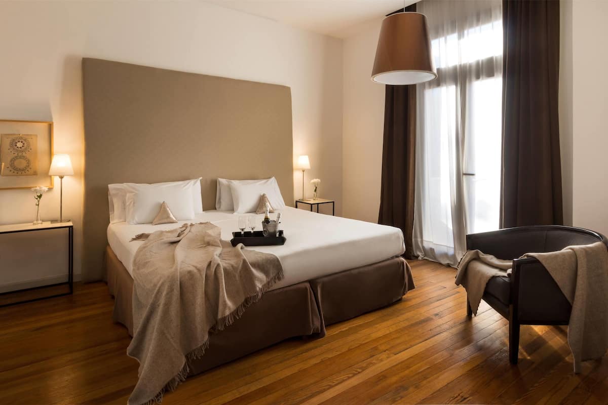 Een netjes ingerichte slaapkamer met een groot bed, nachtkastjes, een stoel en een raam met gordijnen.