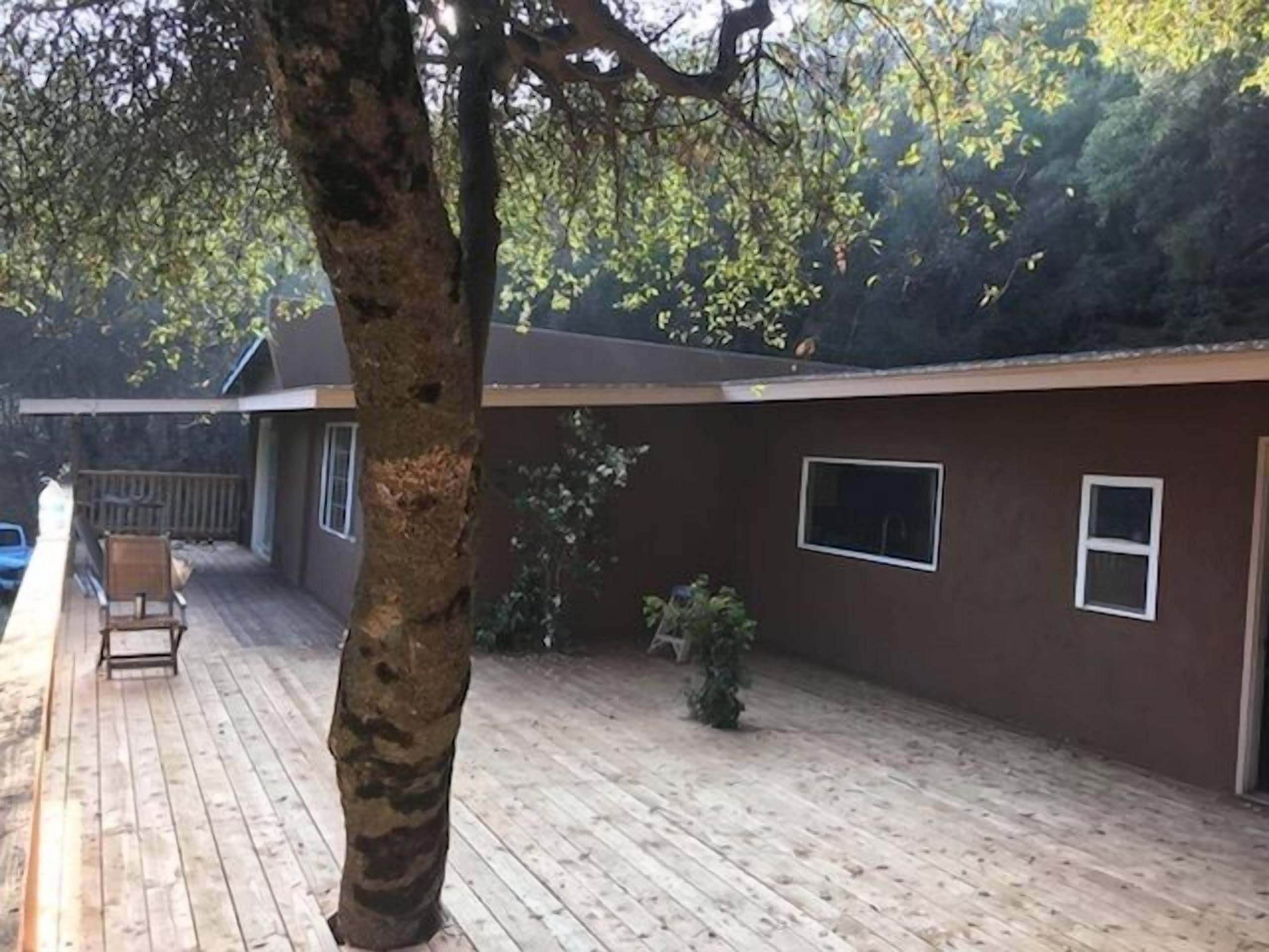 Una casa de un solo piso con una terraza de madera y un gran árbol en primer plano.