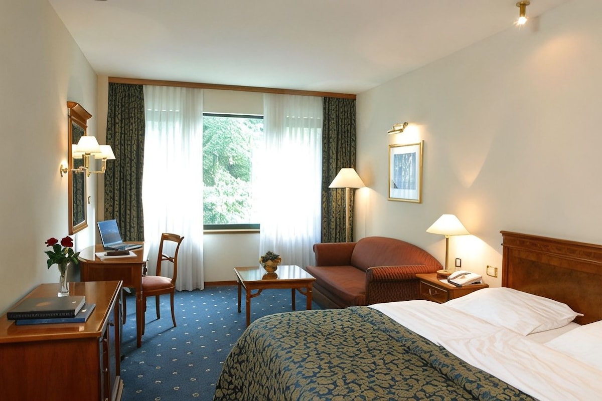 Habitación de hotel con cama, escritorio con una computadora portátil, una silla, un sofá, una mesita con un florero y una ventana con cortinas.