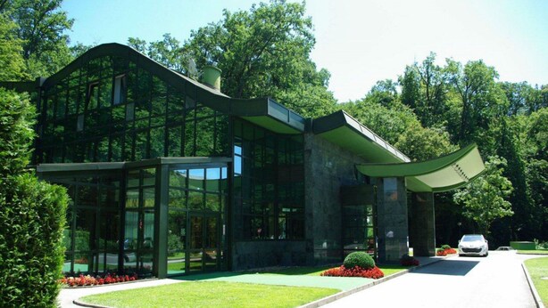 A modern glass building with a curved roof, surrounded by lush greenery and a well-maintained lawn. - Hotel AS (Zagreb)