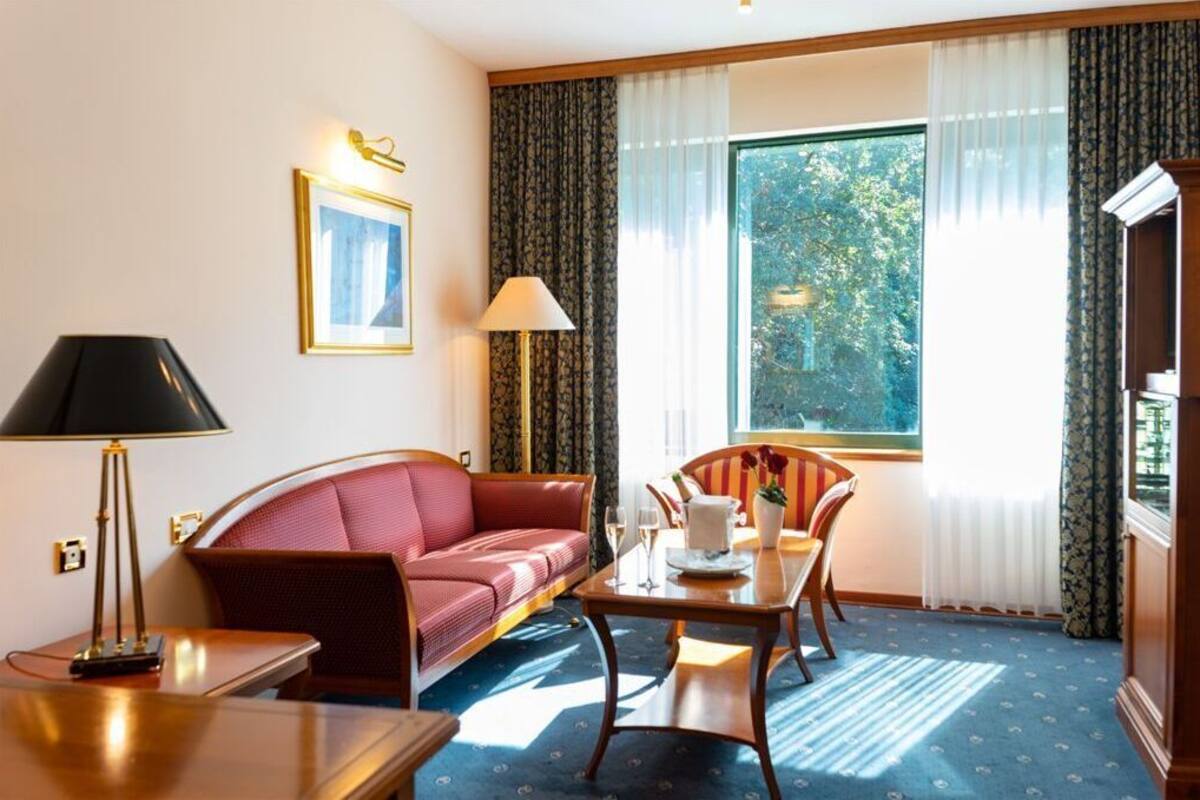 A hotel room with a sofa, a dining table with chairs, a lamp, a picture on the wall, and a window with curtains.