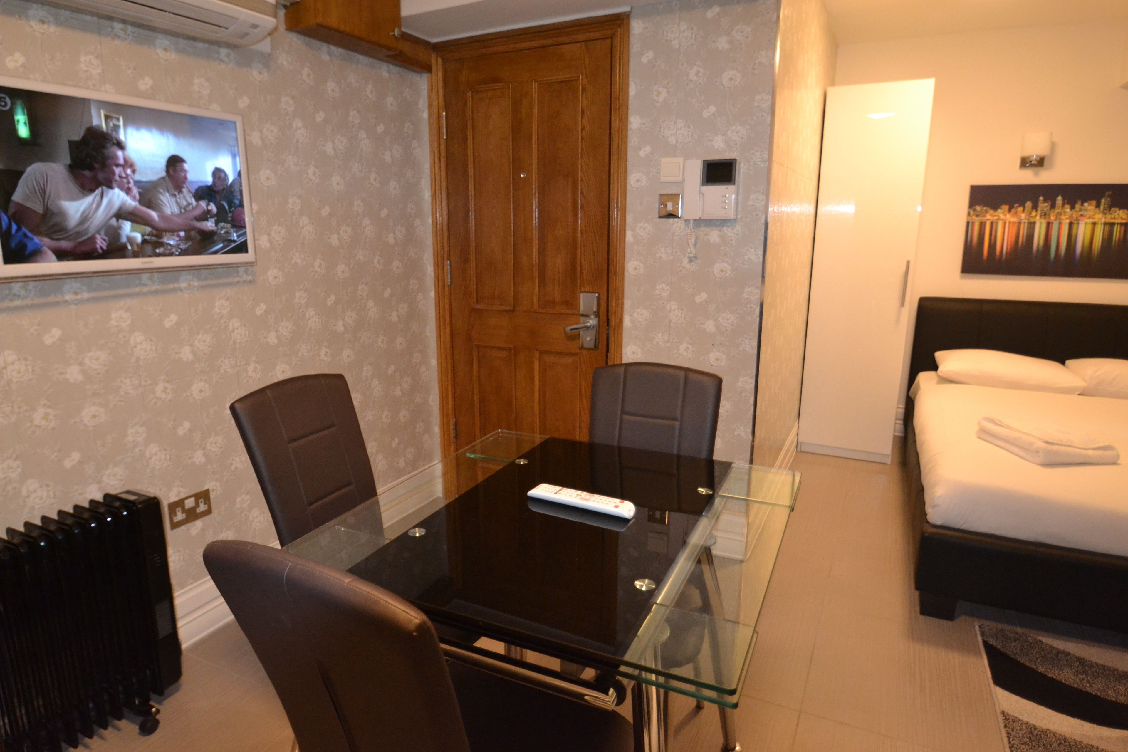Habitación de hotel con mesa de vidrio, dos sillas, cama con ropa blanca, un cuadro en la pared y una puerta de madera.