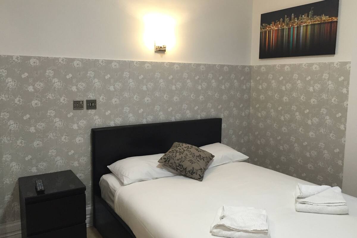 A neatly made bed with white linens, a black headboard, and a floral-patterned wall. There is a small bedside table with a remote control on it, and a painting of a city skyline on the wall above the bed.
