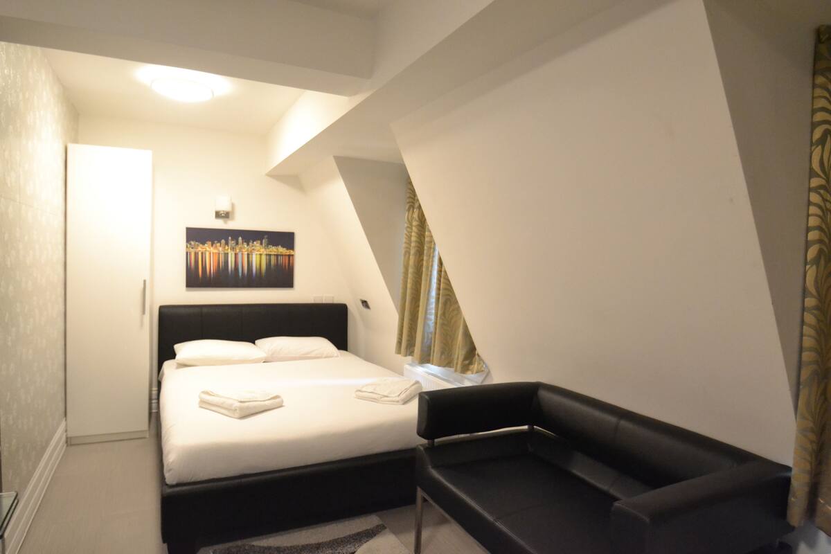 A bedroom with a bed, a black chair, a painting on the wall, and a window with curtains.