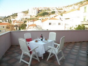 A terrace with a table set for two, white chairs, and a view of a hillside town. - Villa Rosa (Dubrovnik)