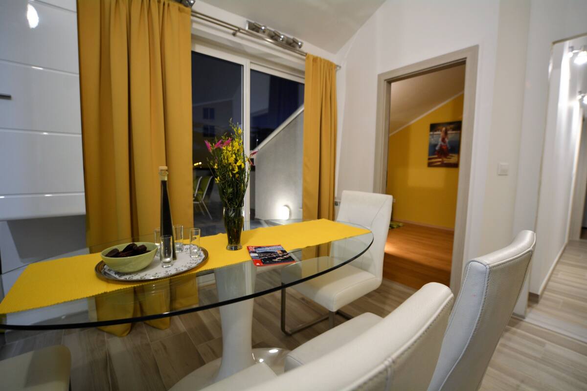 Uma sala de jantar moderna com mesa de vidro, cadeiras brancas, toalha de mesa amarela e uma janela com cortinas.