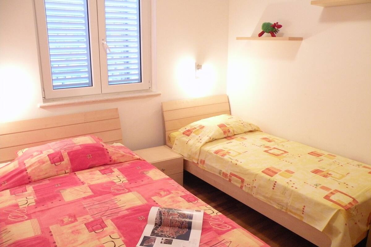 Quarto com duas camas de solteiro, janela com persianas, mesa de cabeceira e uma pequena prateleira com um objeto decorativo.