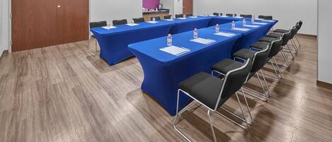Sala de conferência com mesas longas, cadeiras e garrafas de água.