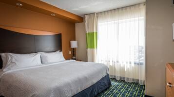 Habitación de hotel con una cama grande, una mesita de noche, una lámpara y vistas a la ciudad a través de cortinas traslúcidas.