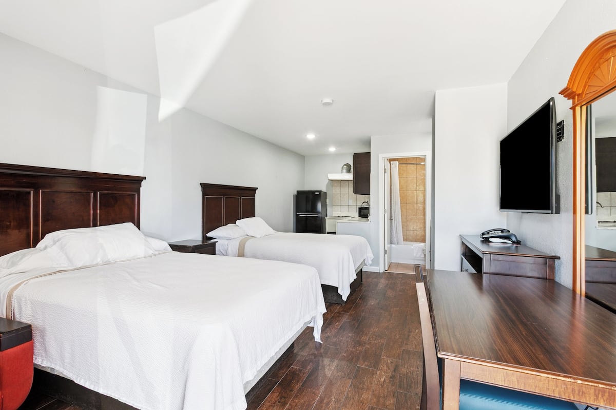 A hotel room with two beds, a wooden desk, a television, and a kitchen area in the background.