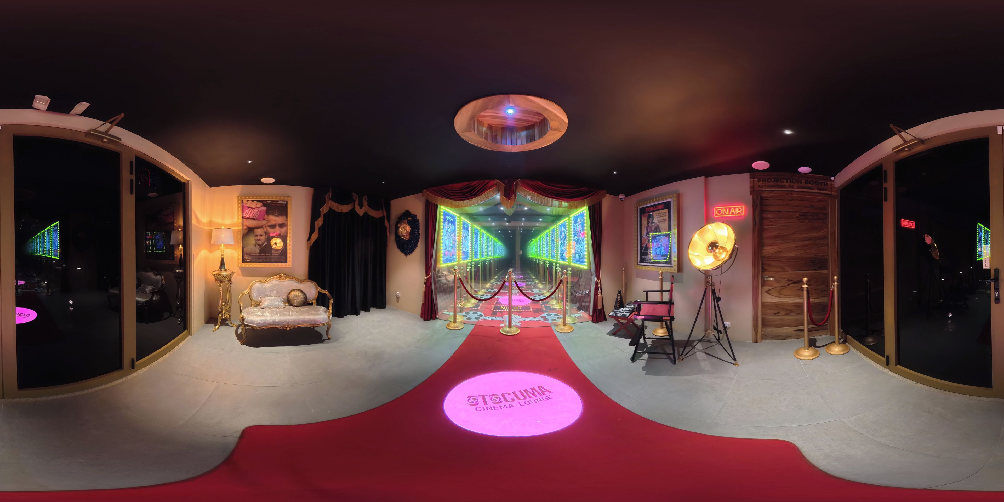 A cinema lounge with a red carpet leading to a seating area, a projection booth, and a wall with illuminated movie posters.
