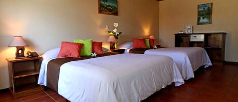 Habitación de hotel con dos camas, cada una con ropa de cama blanca, almohadas rojas y verdes, y una mesita de noche con lámpara y teléfono. Hay un escritorio con cafetera y un cuadro pequeño en la pared.