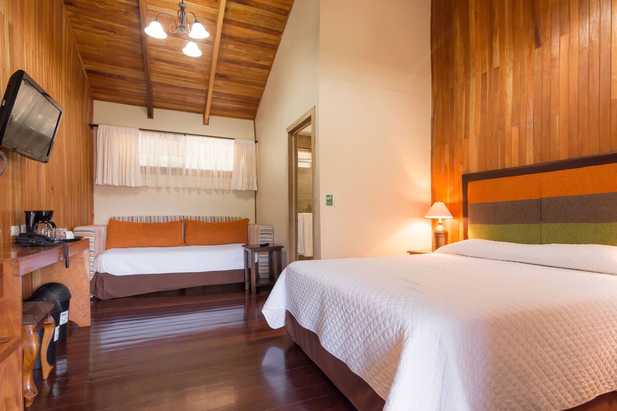 A hotel room with wooden flooring, a large bed with a quilted white duvet, a smaller bed with an orange pillow, a desk with a telephone and a coffee maker, and a television mounted on the wall.