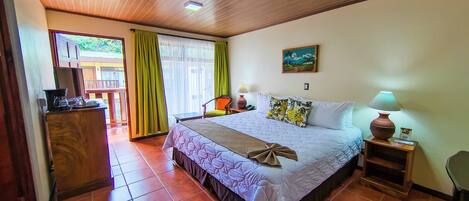 A hotel room with a large bed, a desk with a coffee maker, a chair, a nightstand with a lamp, and a painting on the wall.