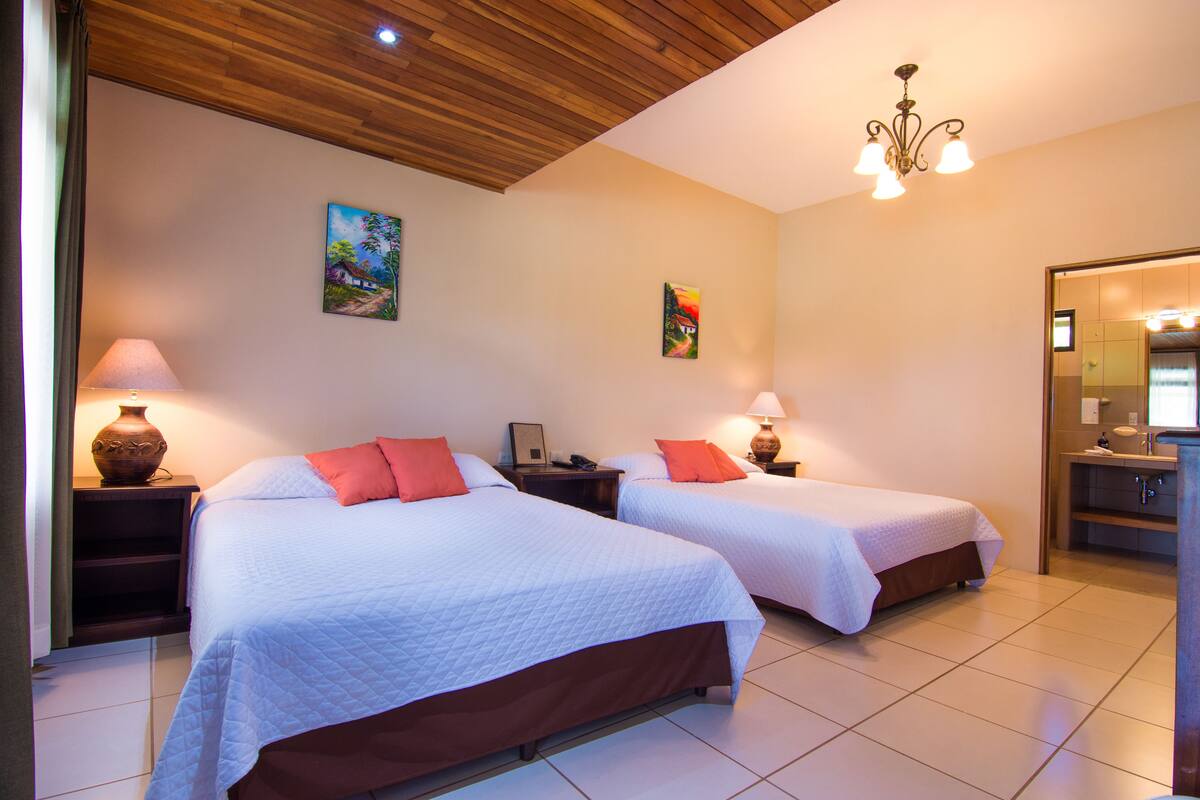 A hotel room with two beds, wooden ceiling, artwork on the walls, and a bathroom visible through an open door.