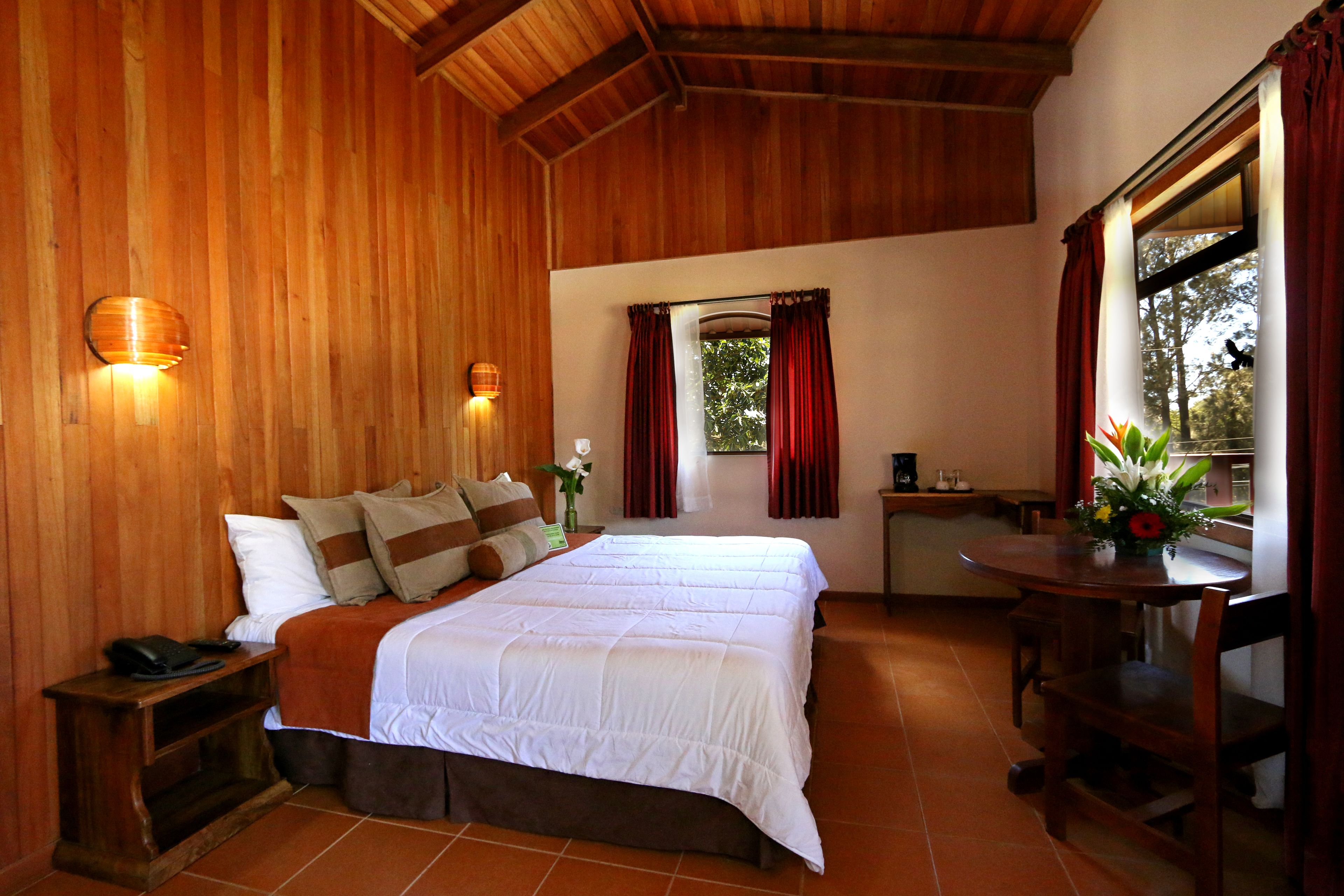 Una habitación con paneles de madera, una cama, una mesa y un ventanal con cortinas.