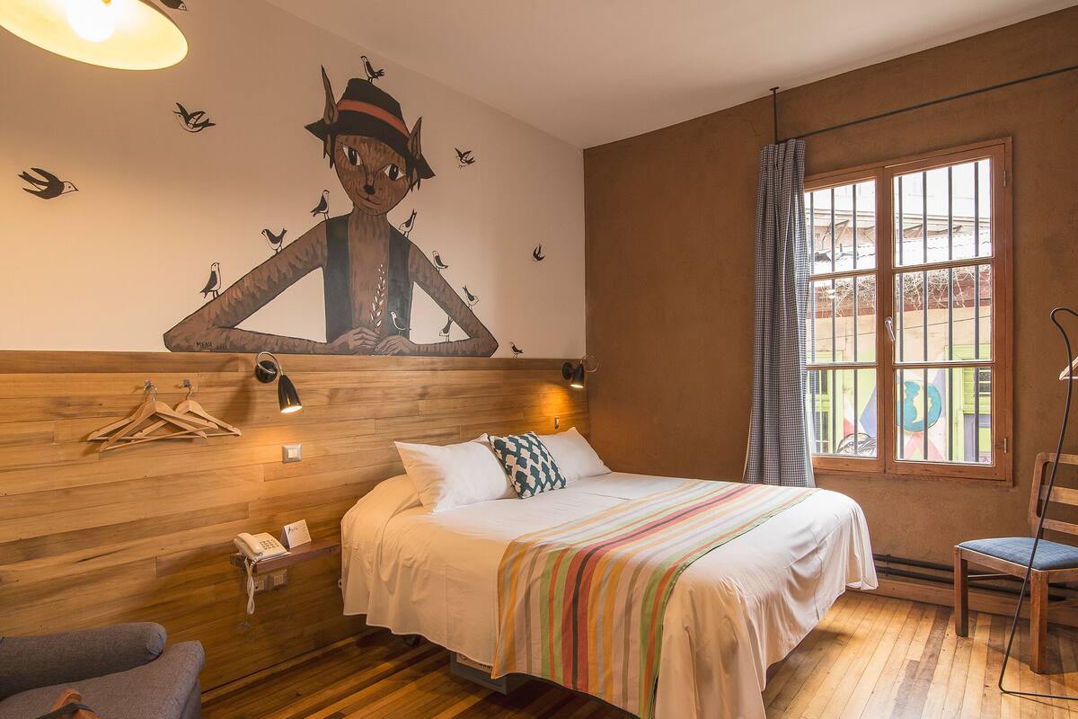 A bedroom with a bed, a wooden headboard, a window with curtains, a chair, a hanging light, and a wall mural.