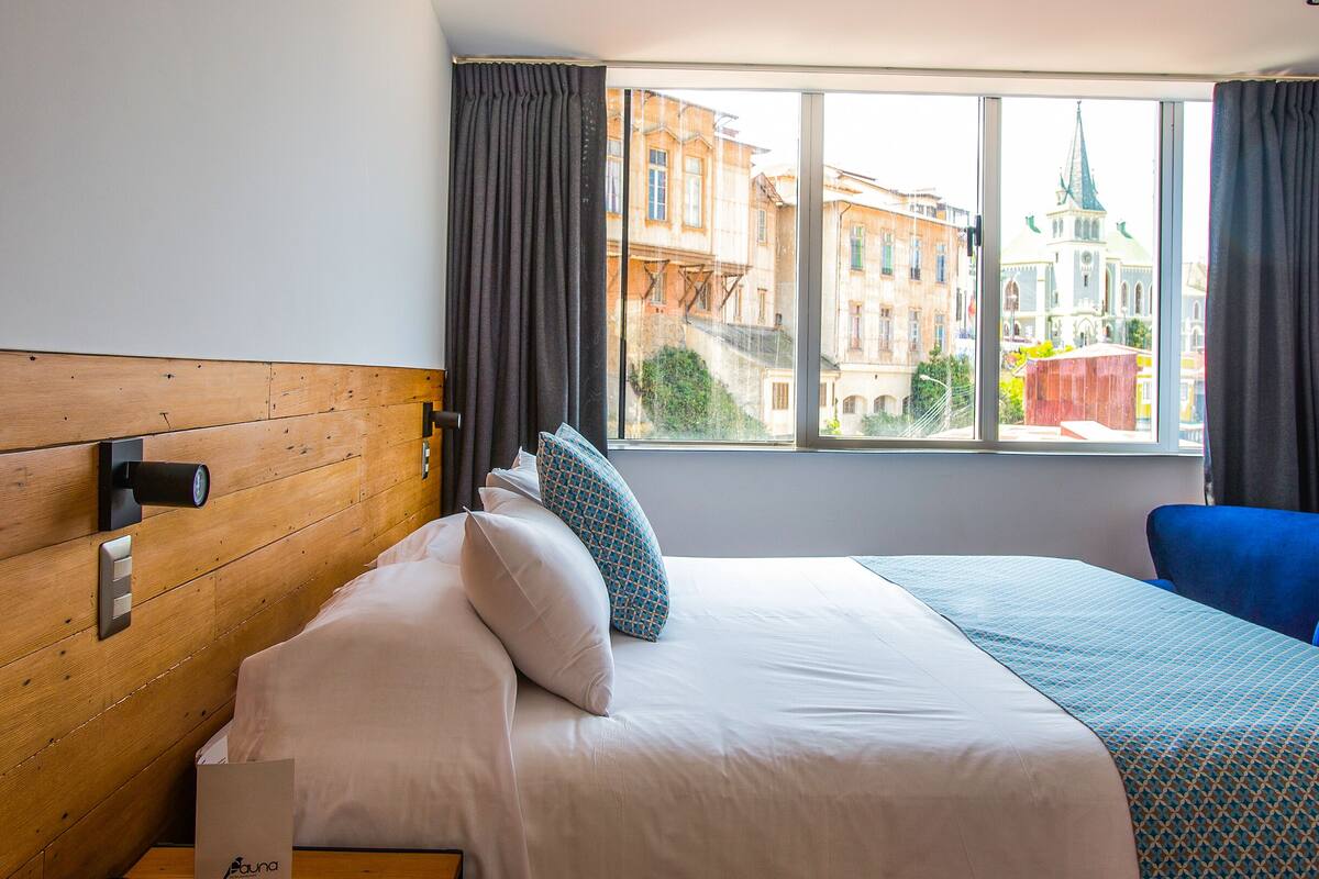A hotel room with a wooden headboard, a bed with white bedding, a blue chair, and a window with a view of buildings and a church spire.