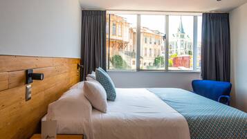A hotel room with a wooden headboard, a bed with white bedding, a blue chair, and a window with a view of buildings and a church spire.
