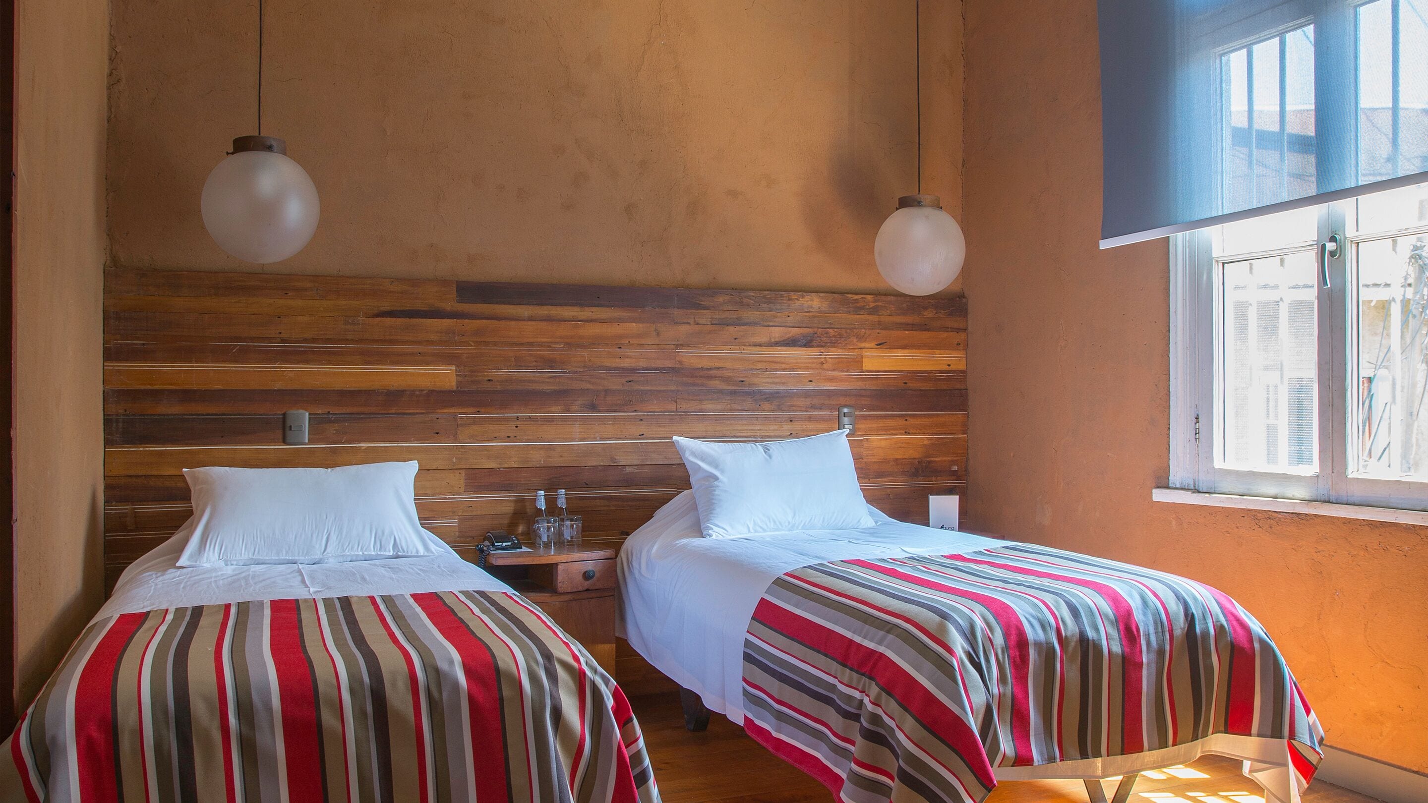 Habitación con dos camas, cabecero de madera, ropa de cama a rayas, lámparas colgantes y ventana con vistas.