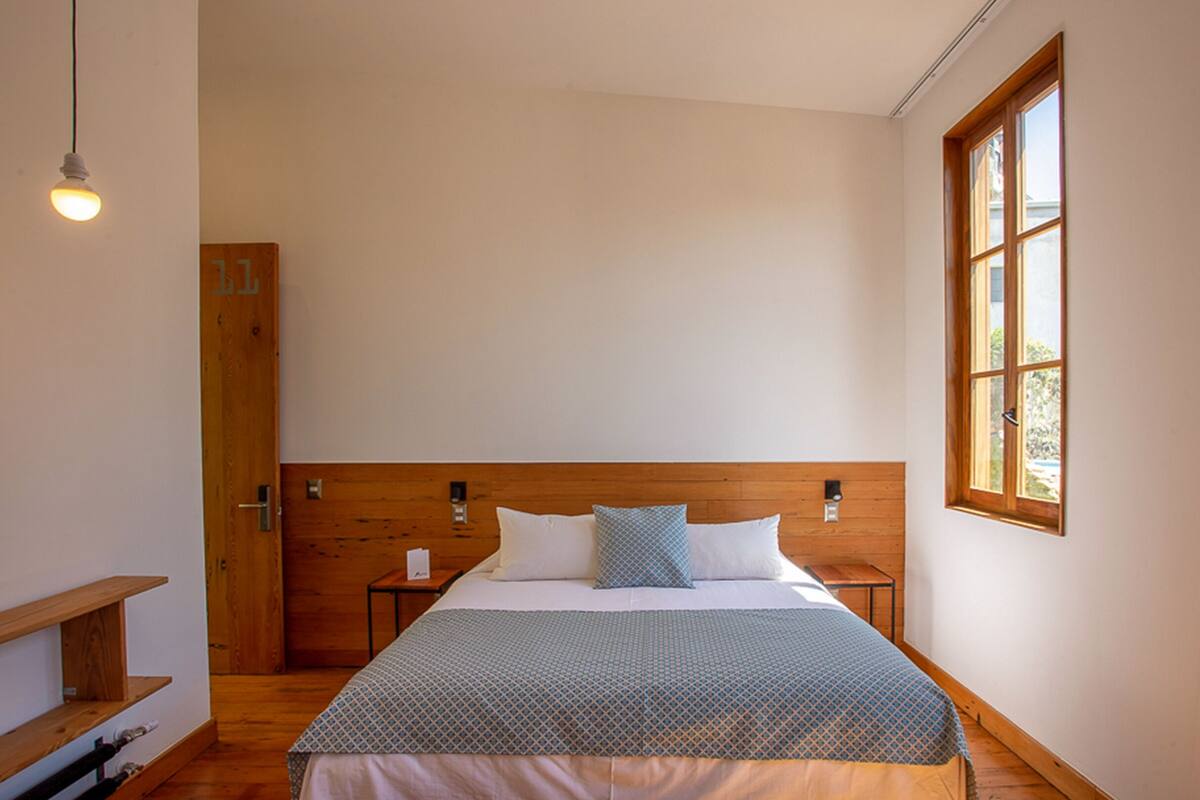 A bedroom with a wooden headboard, a bed with patterned bedding, a nightstand, a bedside table, a hanging light, and a window with a view.
