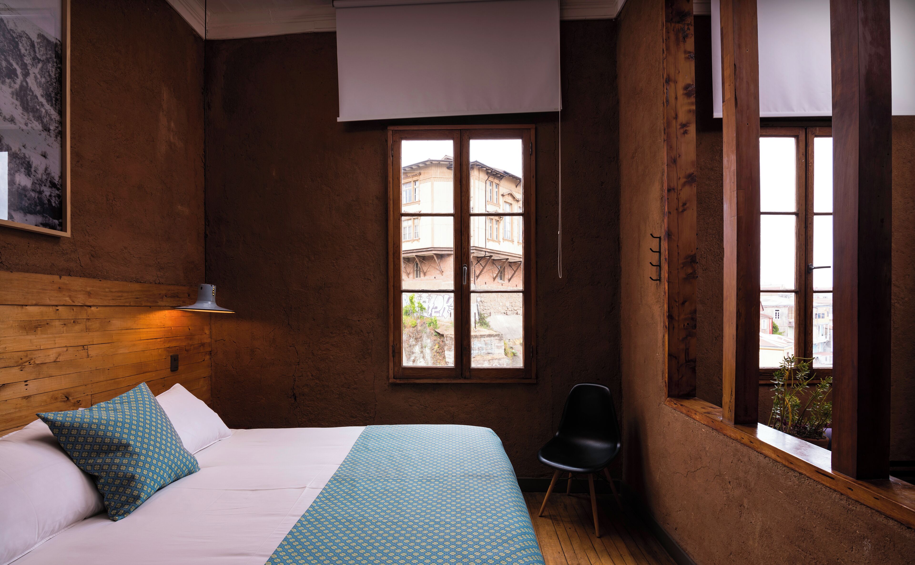 Quarto com cama, uma cadeira, janela com vista para edifícios e parede de madeira.